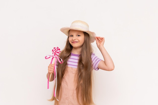 A little girl holds a caramel lollipop on a stick in Baby in a sundress and a summer straw hat on a white isolated background