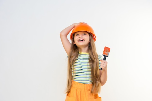 A little girl holds a construction helmet on her head with her hand, and with her other hand holds a paint brush for painting walls in a children's room.