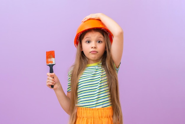 A little girl holds a construction helmet on her head with her hand, and with her other hand holds a paint brush for painting walls in a children's room.