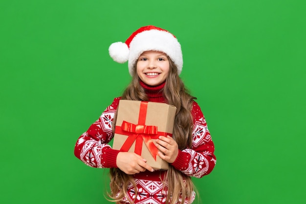 A little girl holds a gift on a green isolated background A child in a Santa Claus hat and a warm sweater holds a gift box