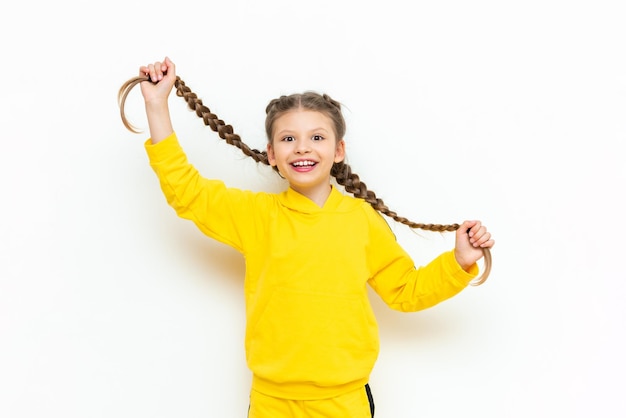 A little girl holds her long hair in braids in her hands and smiles broadly A child in a yellow sports suit on a white isolated background