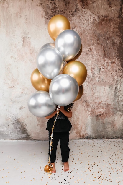 A little girl holds a large bouquet of balloons against the background of a decorative wall holidaynew year birthday golden and silver balloons