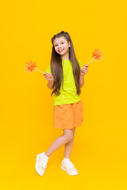 A little girl holds origami flowers on a stick and stands tall Children's creativity A child in shorts and a Tshirt dreams of a sunny summer on a yellow isolated background