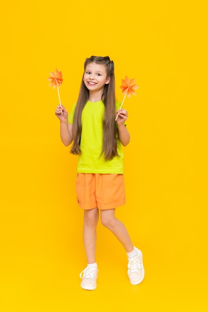 A little girl holds origami flowers on a stick and stands tall Children's creativity A child in shorts and a Tshirt dreams of a sunny summer on a yellow isolated background