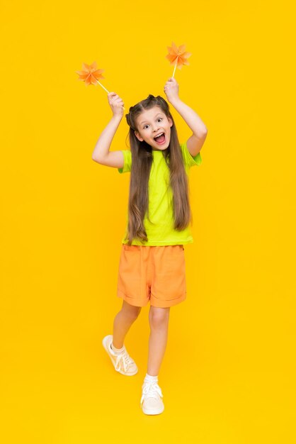 A little girl holds origami flowers on a stick and stands tall Children's creativity A child in shorts and a Tshirt dreams of a sunny summer on a yellow isolated background