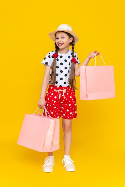 A little girl holds shopping bags A child in a summer hat and shorts is engaged in children's shopping A happy child in full growth on a yellow isolated background