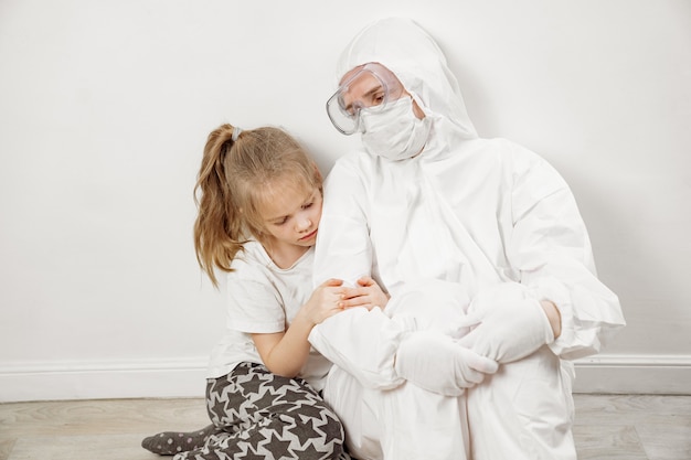 A little girl hugs a doctor in a white protective suit, mask, glasses and gloves. red zone. Baby patient. Gratitude. medicine during a pandemic. mom is a doctor