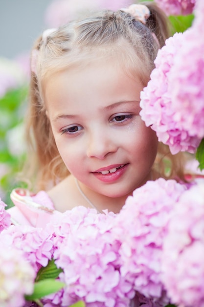 Little girl is in bushes of hydrangea flowers in sunset garden.