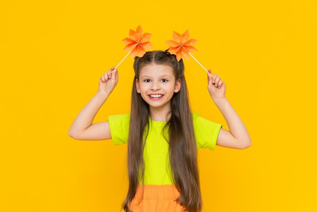 A little girl is playing with paper flowers on sticks Origami mill Crafts made of paper Children's creativity A beautiful child on a yellow isolated background