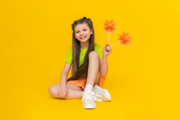 A little girl is playing with paper flowers on sticks An origami mill Happy kid holds paper crafts Children's creativity A beautiful child is sitting on a yellow isolated background