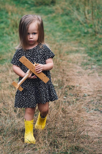 A little girl is playing with a wooden airplane in a village in a field Children's happiness happy childhood concept A beautiful blonde girl in a dress and boots looks thoughtfully