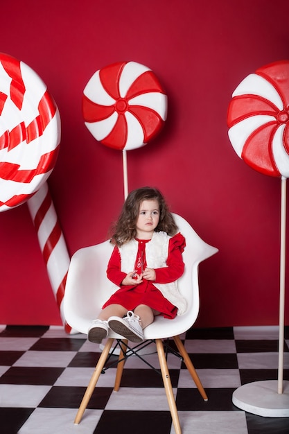 Little girl is sitting on chair with big christmas candies on red background