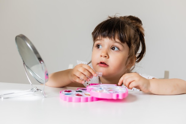 Little girl is sitting at the dressing table in front of the mirror and applying makeup like Mom