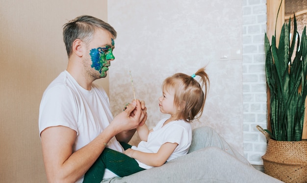 A little girl is sitting on her father's lap painting his face with a brush of oil paints. body art art