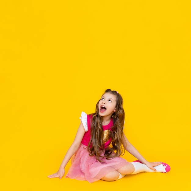 A little girl is sitting and looking up at your advertisement on a yellow isolated background A child with long wavy hair in a pink dress is smiling broadly