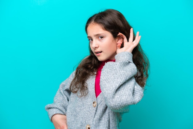 Photo little girl isolated on blue background listening to something by putting hand on the ear