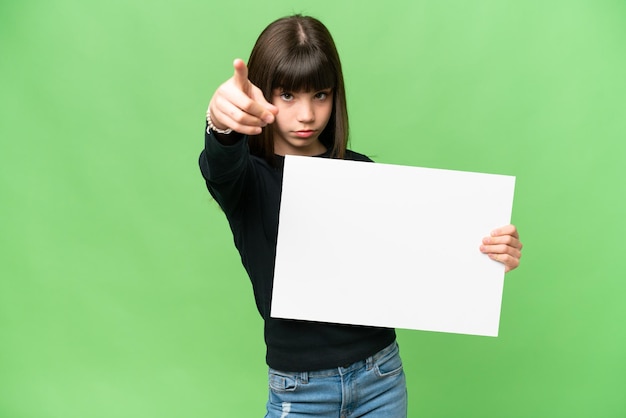 Little girl over isolated chroma key background holding an empty placard and pointing to the front