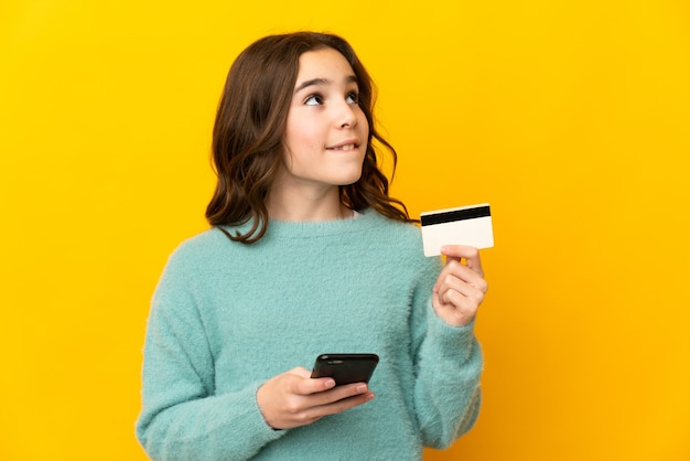 Little girl isolated on yellow wall buying with the mobile with a credit card while thinking