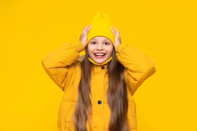 A little girl in a jacket and hat holds her head and smiles happily A child in autumn fashionable orange clothes Dress warmly in autumn Yellow isolated background