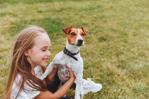 A little girl kisses and hugs her Jack Russell terrier dog in the park Love between the owner and the dog a child is holding a dog in his arms