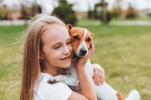 A little girl kisses and hugs her Jack Russell terrier dog in the park Love between the owner and the dog a child is holding a dog in his arms