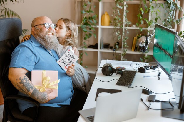 Little girl kisses smiling bearded dad with gift box and greeting card in home office