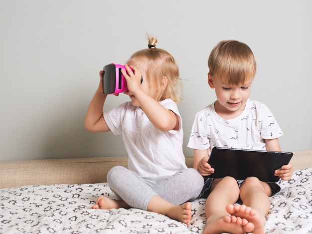 Little girl looking in virtual reality glasses and boy with touch pad on bed. Scandinavian interior