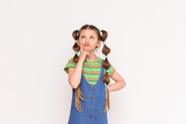 A little girl looks up thoughtfully with funny ponytails and a summer denim sundress on a white isolated background