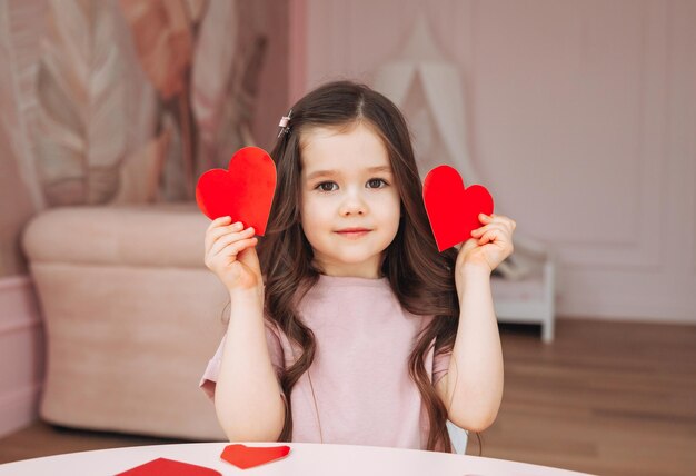 A little girl makes Valentine's Day cards using colored paper scissors and pencil sitting at a table