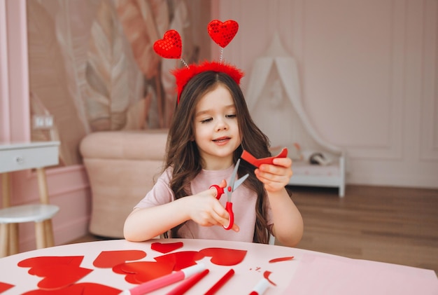 A little girl makes Valentine's Day cards using colored paper scissors and pencil sitting at a table