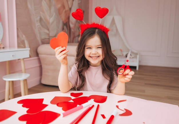 A little girl makes Valentine's Day cards using colored paper scissors and pencil sitting at a table