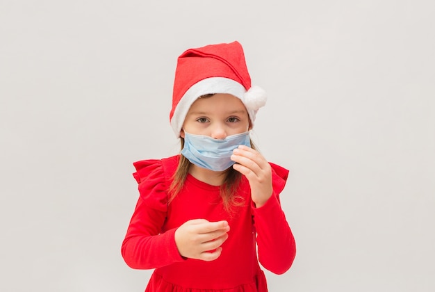 Little girl in a medical mask in a red cap on a white wall with space for text