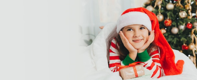 Little girl in a New Year's red hat with a Christmas gift She lies on the bed hugging a box against the background of a Christmas tree