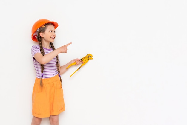 A little girl in an orange construction helmet holds a monkey wrench and points at your advertisement on a white isolated background Copy space