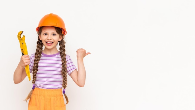 A little girl in an orange construction helmet holds a monkey wrench and points at your advertisement on a white isolated background Copy space