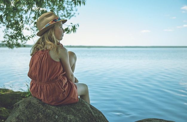 A little girl in an orange dress and hat is sitting on a stone on the shore of a lake and looking dreamily into the distance on a summer day Tint image