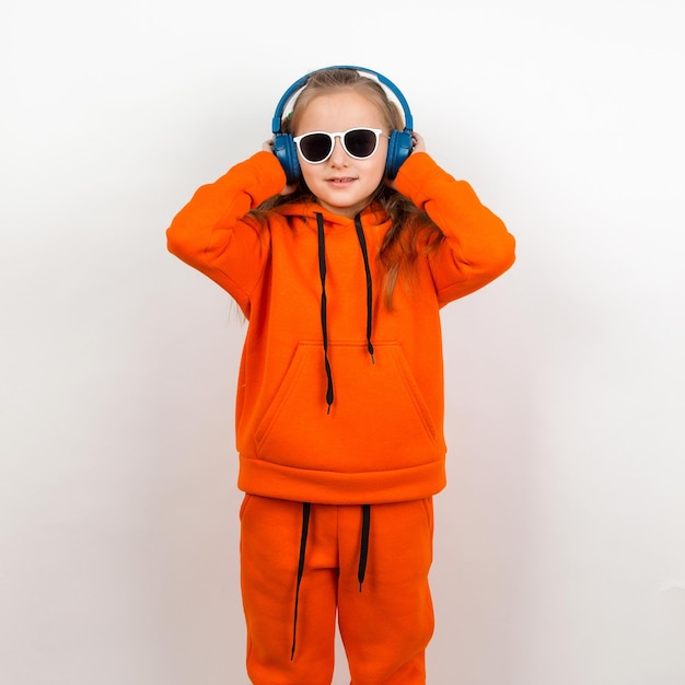 A little girl in an orange hoody sunglasses and blue headphones listens to music Portrait on a white background