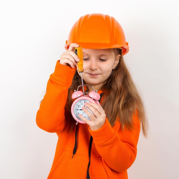 A little girl in an orange suit and an orange construction helmet Fixes the alarm clock with screwdriver White background