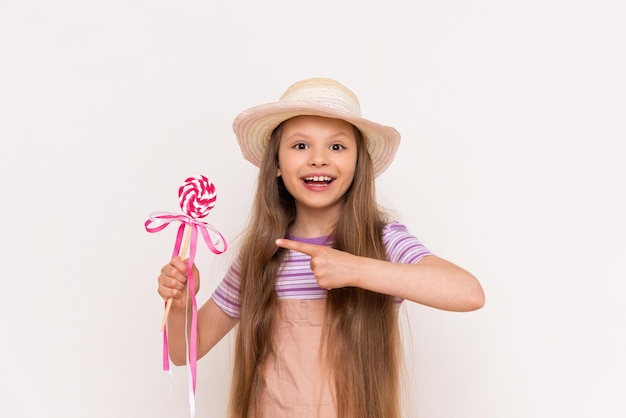 A little girl in overalls and a summer hat points to a caramel lollipop on a white isolated background Sweets for children