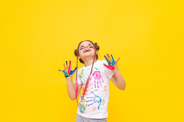 A little girl painted with paints looks at your advertisement on a yellow isolated background The art of painting with paints for children Art courses for schoolchildren Copy space