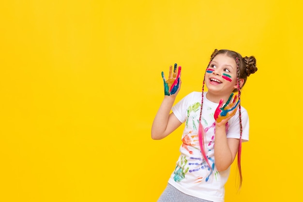 A little girl painted with paints looks at your advertisement on a yellow isolated background The art of painting with paints for children Art courses for schoolchildren Copy space