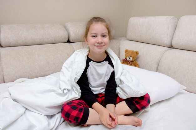 Little girl in pajamas sits in bed covered with a blanket and smiles