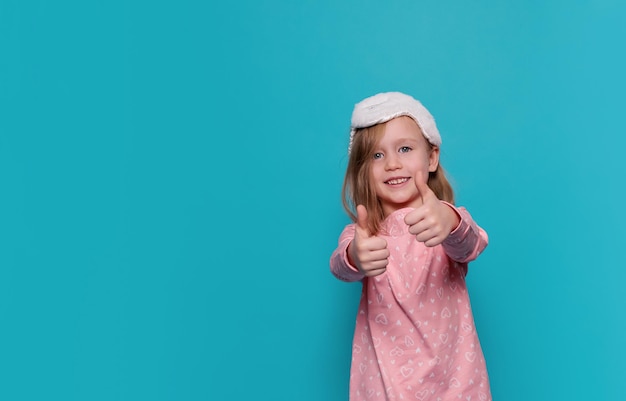 A little girl in pajamas and a sleep mask shows a like on a blue background copy space