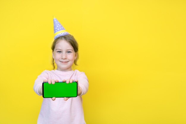 Little girl in a paper cap on outstretched arms shows a phone with a green screen, copy space