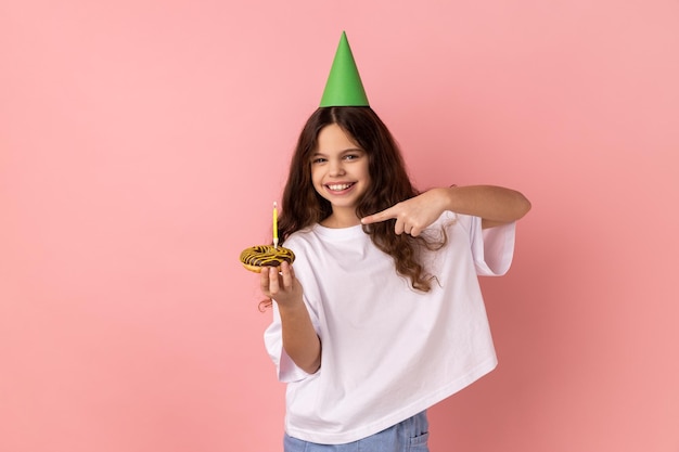 Little girl in party cone on head pointing at cake with candle and looking at camera with smile