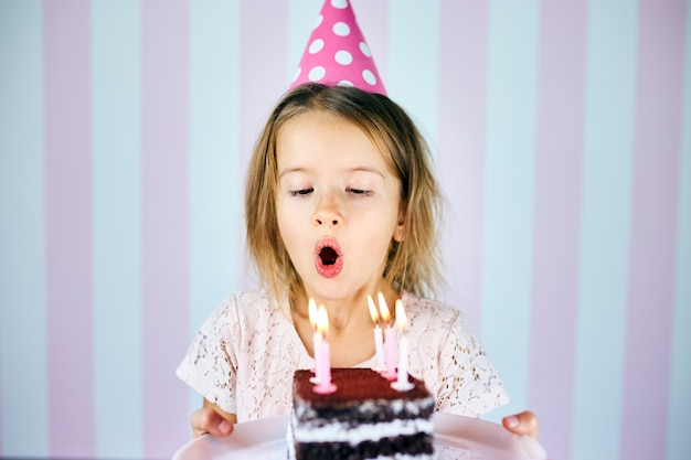 Little girl in pink cap blowing out candles on a birthday chocolate cake