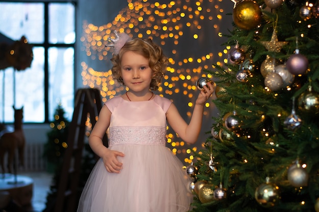 a little girl in a pink dress and a crown near the Christmas tree with gifts.