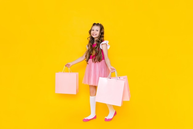 A little girl in a pink dress holds large shopping bags on a yellow isolated background Spring shopping in the store big promotions on goods