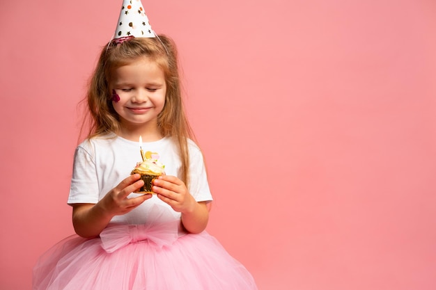 A little girl in a pink dress with a birthday cake on a pink background.