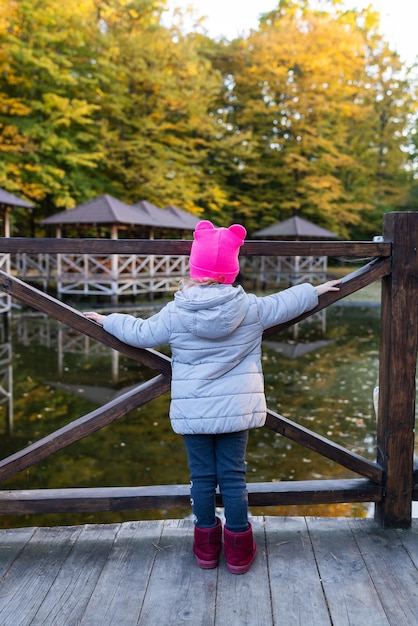 A little girl in a pink hat stands with her back near the pond and enjoys a warm autumn day